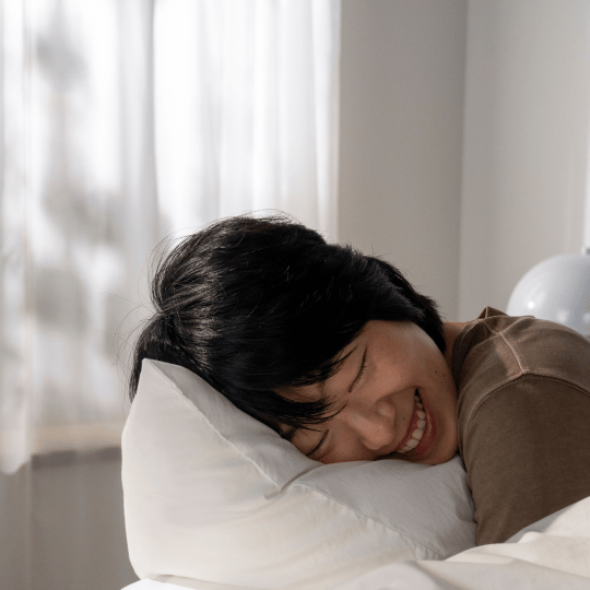 A person with dark hair smiles with closed eyes while lying on a white pillow.