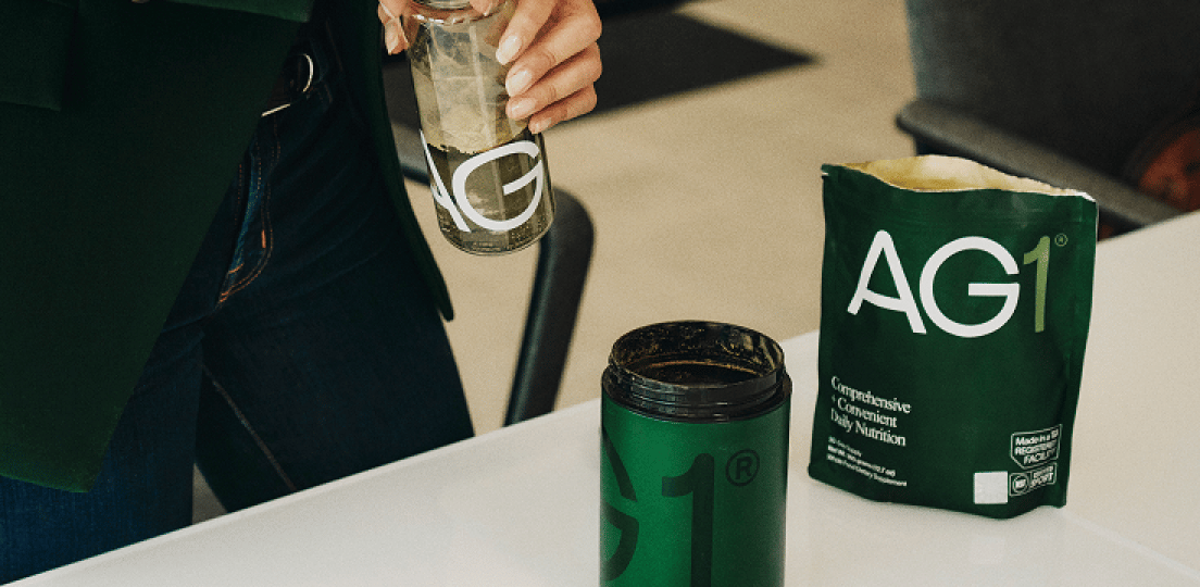 A hand pours green liquid from a bottle into a glass with ice held by another hand.