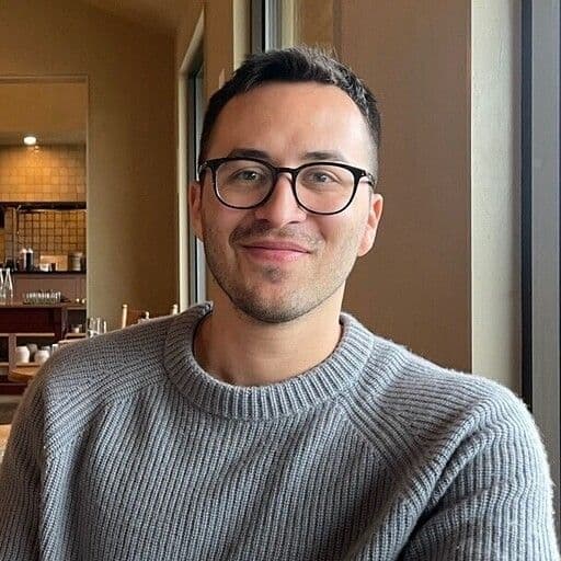 A man with glasses and a grey sweater smiles at the camera.