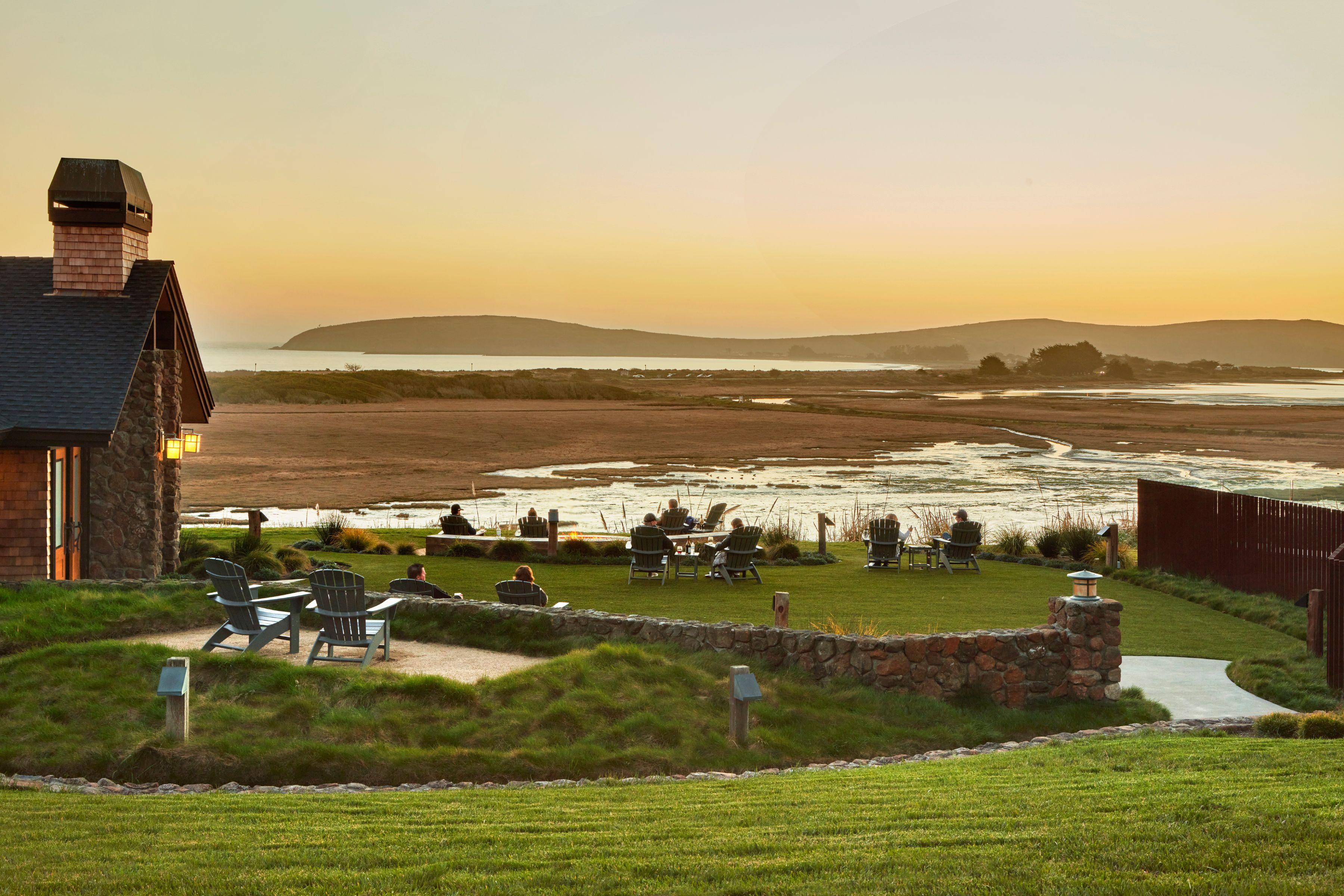 Rustic lodge and grassy lawn with people relaxing at sunset, overlooking a marshy bay with distant hills.
