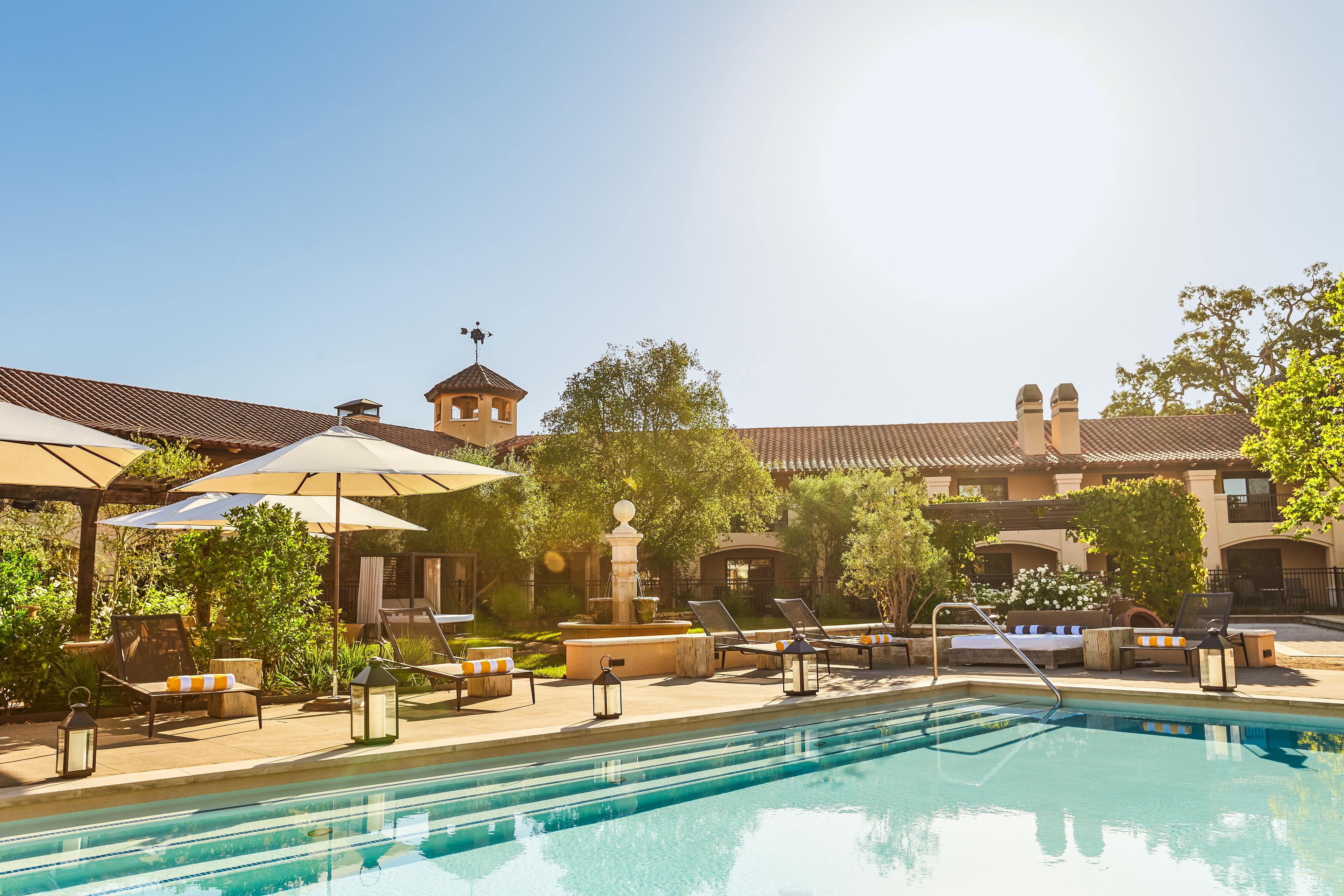 A hotel swimming pool area on a sunny day, with lounge chairs, umbrellas, and a building with a tower in the background.