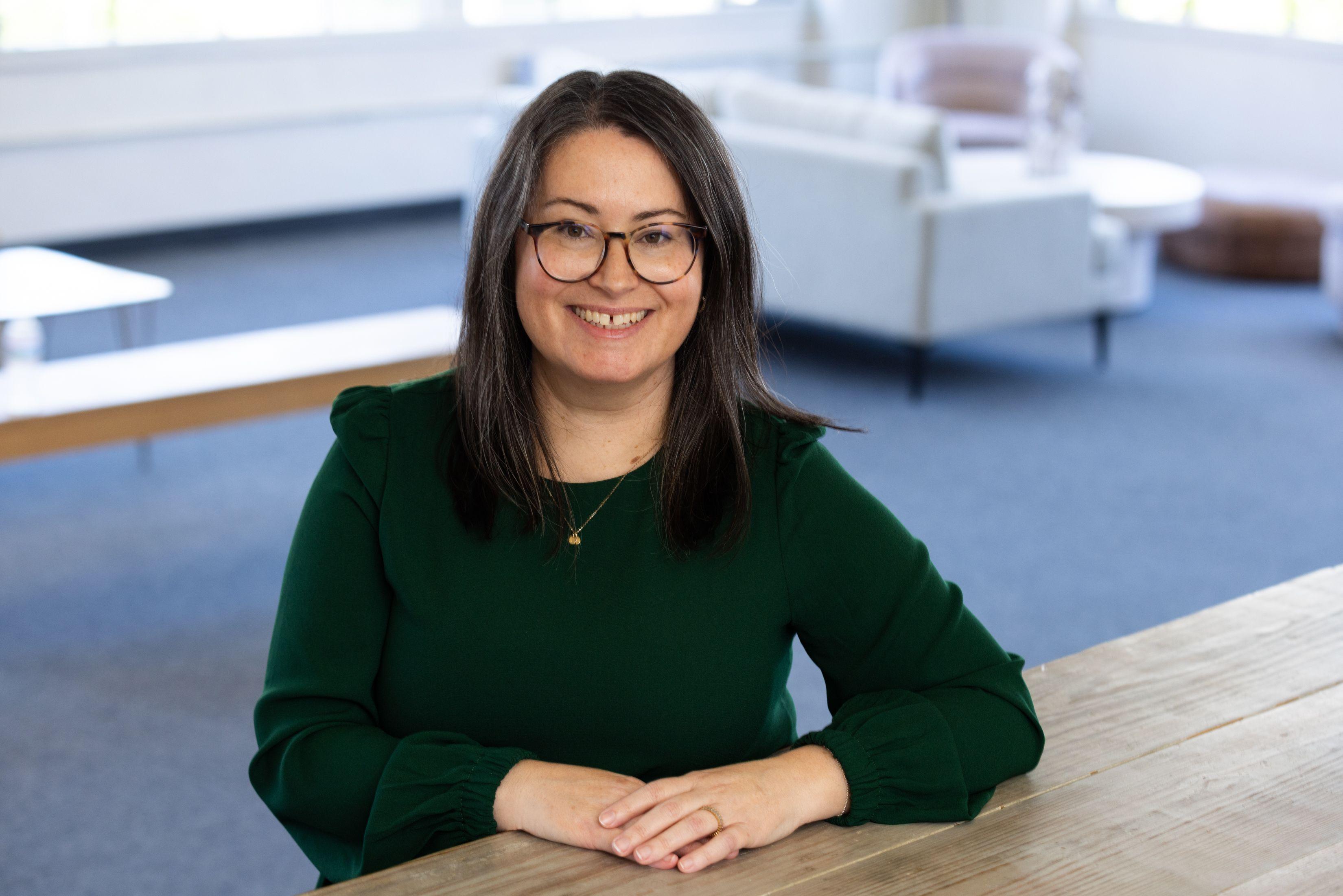 A smiling woman with dark hair and glasses in a green top sits at a wooden table in a bright office.
