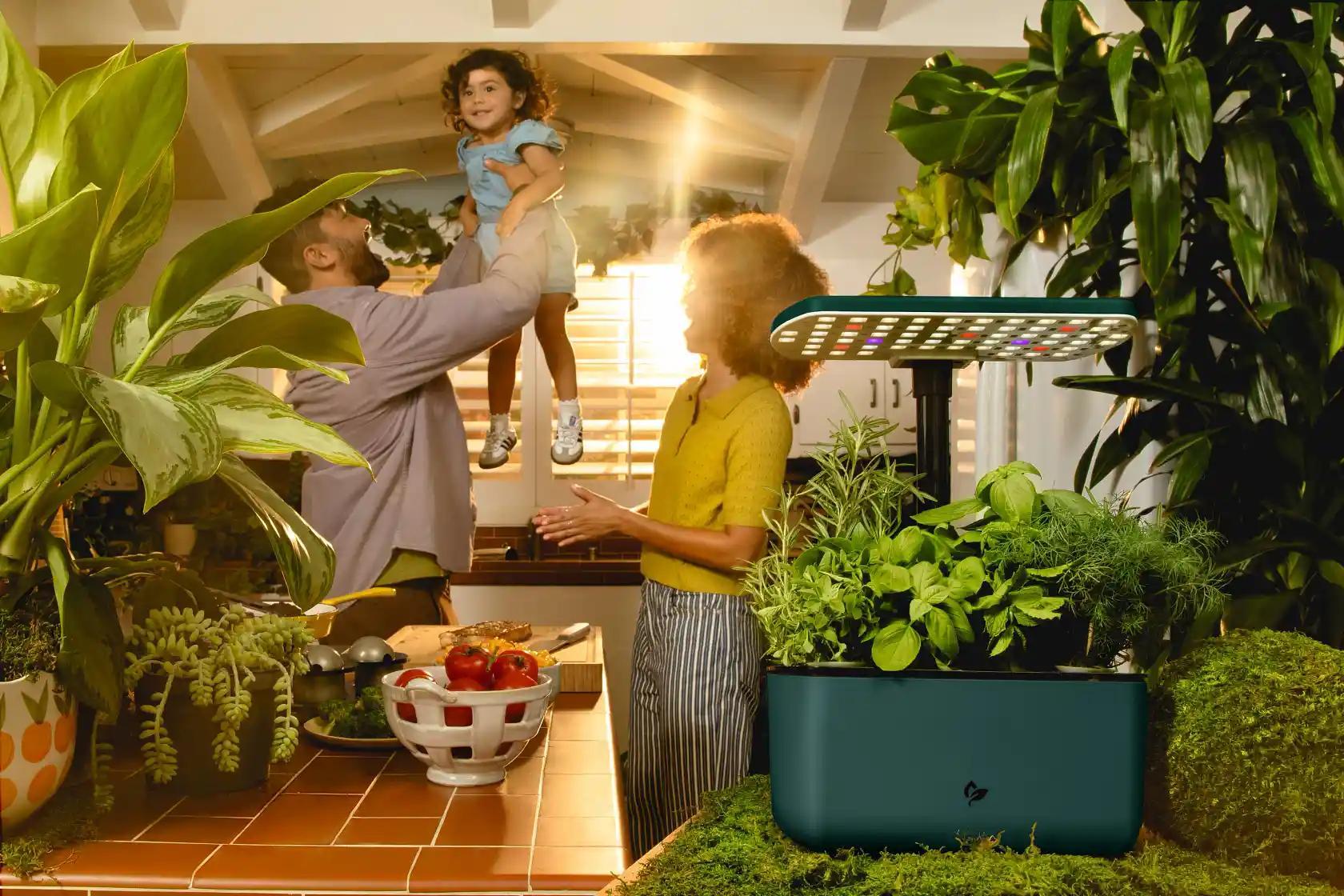 An indoor smart garden with herbs and a grow light sits in a bright kitchen where a father lifts his child as a woman looks on