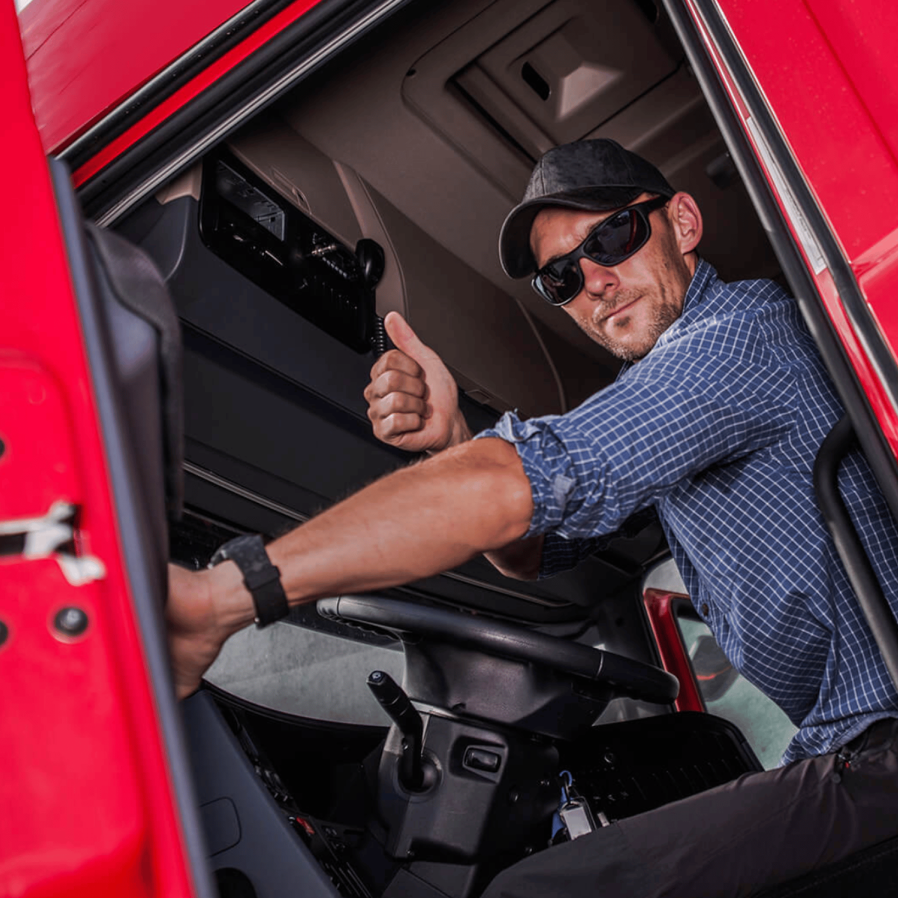 A man in sunglasses and a cap sits in a red truck and gives a thumbs-up.