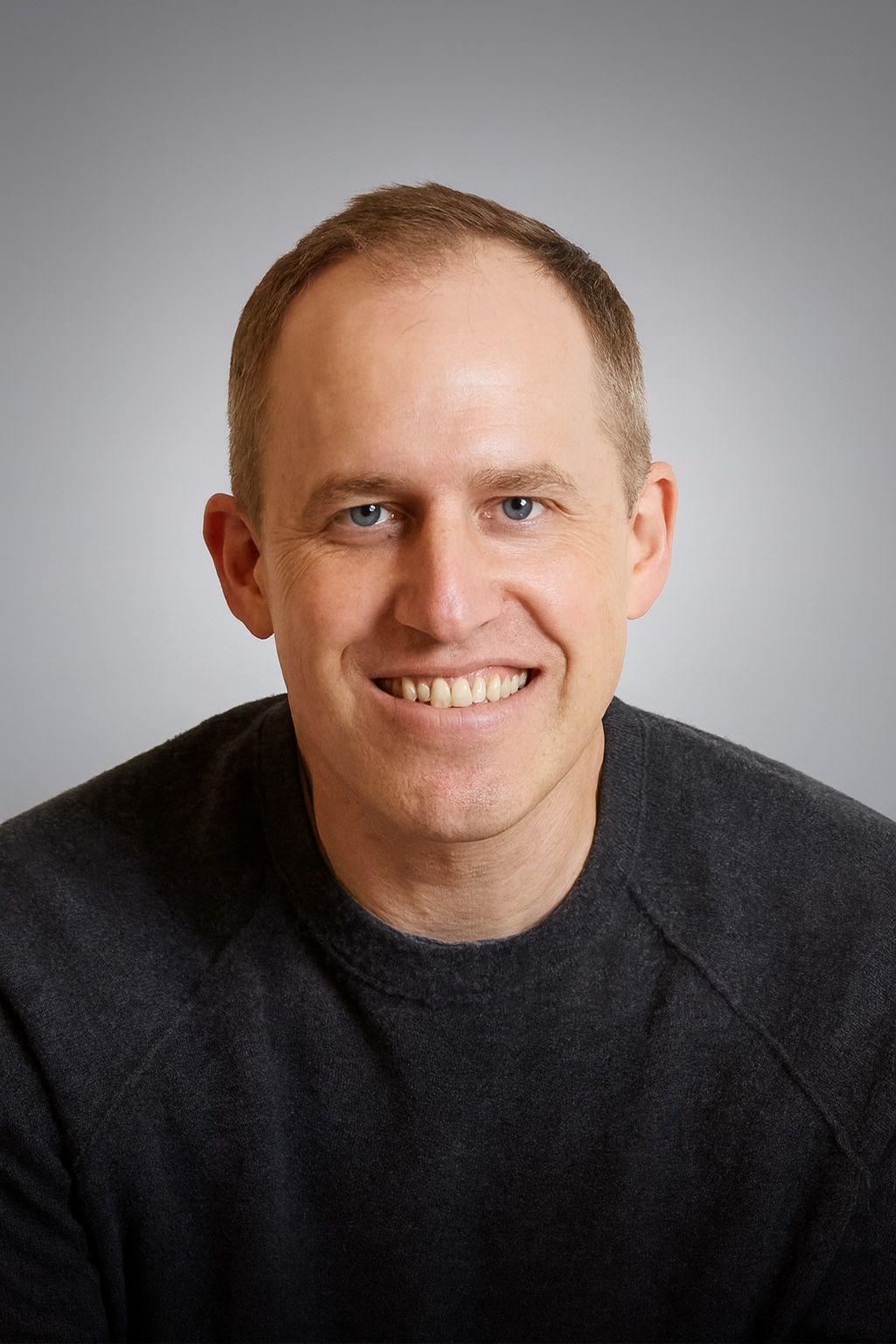 A smiling man with blue eyes and light brown hair, wearing a dark gray sweater.