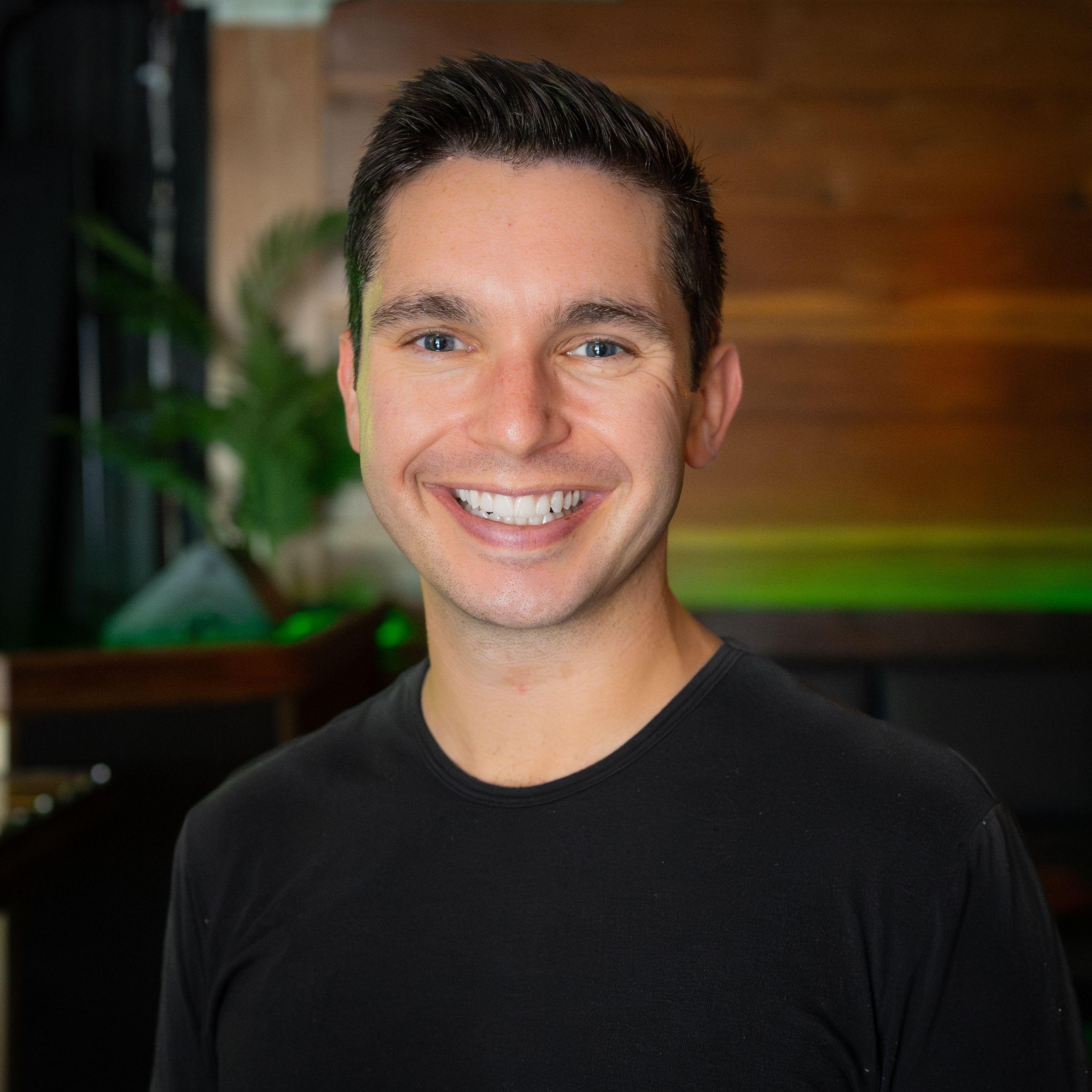 A smiling man with dark hair and blue eyes wearing a black t-shirt.